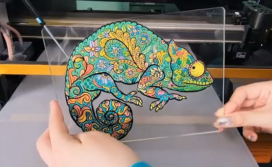Hands holding a clear acrylic panel with a colorful chameleon print, intricate floral design, printed by a flatbed printer.
